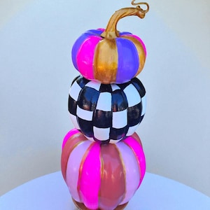 May include: A vertical stack of three decorative pumpkins. The top pumpkin features alternating vertical stripes of neon pink, purple, and metallic gold. The middle pumpkin has a black and white checkerboard pattern. The bottom pumpkin has vertical stripes of neon pink, light pink, and metallic gold.