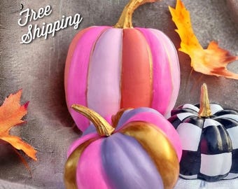May include: A collection of decorative pumpkins in various colors. One pumpkin is pink, orange, and light pink with a gold stem. Another is pink and purple with a gold stem. A third is black and white checkered. Autumn leaves are scattered around the pumpkins.