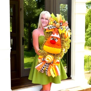 May include: A festive Halloween wreath featuring a large candy corn design with a smiling face, surrounded by yellow and orange ribbons, bows, and decorative elements. The wreath is designed with a mix of textures and colors, perfect for seasonal decor.