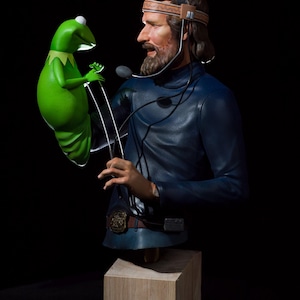 Könnte beinhalten: Eine grüne Froschpuppe mit einem breiten Maul und großen Augen wird von einem Mann in einem blauen Overall und einem Headset gehalten. Der Mann hat einen Bart und schaut die Puppe an. Die Puppe steht auf einem Holzsockel.