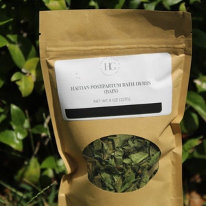 May include: A brown paper bag with a clear window displaying dried green herbs. The bag has a white label with the text "HAITIAN POSTPARTUM BATH HERBS (BAIN)" and "NET WT 8 OZ (227g)". The background is blurred green foliage.