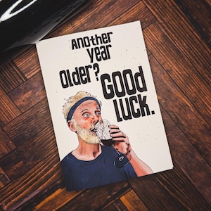 May include: A greeting card with the text "Another Year Older? Good Luck." features an illustration of a man drinking wine. A dark wine bottle is visible in the background. The card is on a wooden surface.