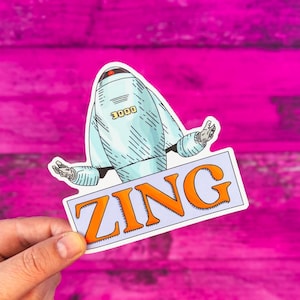 May include: A die-cut sticker featuring a light blue robot with the number 3000 printed on its chest. Below the robot, a rectangular light purple banner displays the word ZING in bold orange capital letters with a dark outline.