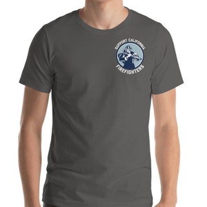 Puede incluir: Una camiseta gris oscuro con un gráfico circular blanco que muestra a un bombero sosteniendo una manguera y el texto "Support California Firefighters".