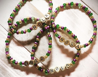 Peut inclure: Trois bracelets de perles circulaires présentant un motif répétitif de perles dorées métalliques, vertes translucides et violettes opaques. Chaque bracelet comprend quatre perles dorées métalliques rondes avec le texte NOLA en noir.