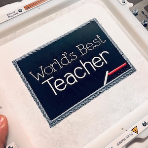 May include: A black rectangular patch with the words "World's Best Teacher" embroidered in white. A red and white line is below the word "Teacher". The patch is on a white surface, possibly fabric, within an embroidery machine frame.