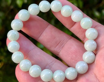 Pode incluir: Uma pulseira de contas de jade verde claro. As contas redondas e polidas têm uma superfície lisa e brilhante. A pulseira é mostrada em uma mão. Cada conta tem cerca de 1 cm de diâmetro.
