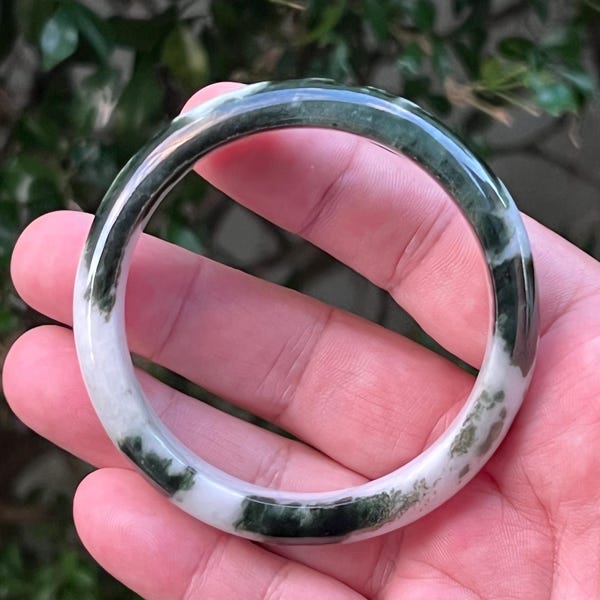 May include: A jade bangle bracelet with a mix of white and dark green colors. The bracelet is circular and appears to be made of a polished stone. The green hues are mottled throughout the white base, creating a natural, organic pattern. The bracelet is held in a hand.