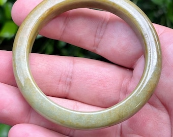 May include: A smooth, circular bangle bracelet crafted from polished stone with a translucent quality. The bangle features a variegated color palette of muted olive green, earthy yellow, and soft brown tones throughout its solid, rounded surface.