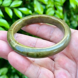 May include: A jade bangle bracelet in shades of green and brown. The smooth, polished surface reflects light, highlighting the natural variations in colour. The bracelet is held in a hand, with a blurred green background.