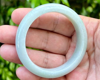 May include: A smooth, circular bangle bracelet crafted from a solid piece of pale, translucent light green stone. The bangle features a polished, rounded surface with subtle variations in color and texture throughout the material.