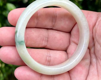 May include: A smooth, circular bangle bracelet crafted from translucent off-white stone with subtle, pale green inclusions. The polished surface of the solid, continuous band reflects light, showcasing a natural, semi-opaque appearance throughout.