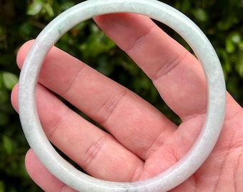 May include: A smooth, circular bangle bracelet crafted from pale, translucent green jade stone. The bangle features a polished, rounded surface with subtle variations in color, ranging from milky white to soft, light green hues throughout the stone.