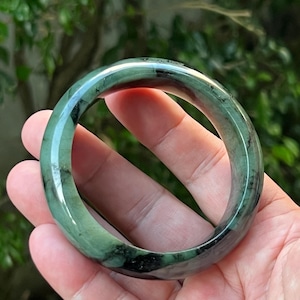 May include: A jade bangle bracelet with a smooth, polished surface. The bracelet is a deep green color with black veining throughout. The bangle is round and appears to be made of a solid piece of jade. The background is blurred, with green foliage.