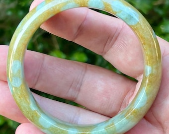 May include: A smooth, polished bangle bracelet featuring a mottled pattern of translucent pale mint green and warm golden yellow tones. The circular jewelry piece has a rounded profile and a glossy, reflective surface throughout its entire circumference.