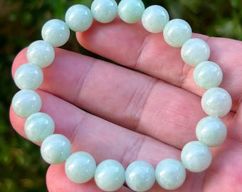 Pode incluir: Uma pulseira de contas verde claro. A pulseira é feita de contas redondas e polidas. As contas têm uma aparência macia e translúcida. A pulseira é mostrada na mão de uma pessoa. O fundo é folhagem verde desfocada.
