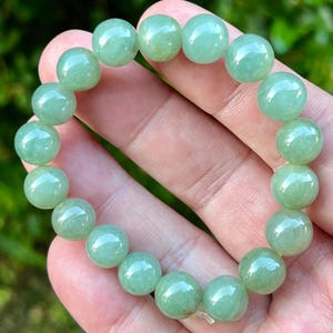 May include: A light green beaded bracelet. The bracelet is made of round, polished beads. The beads have a translucent quality and a subtle variation in color. The bracelet is shown against a blurred background.