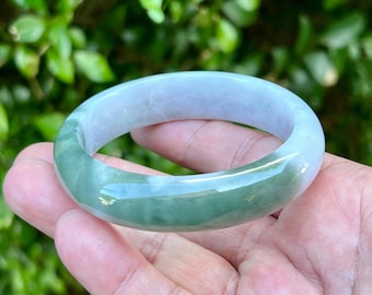 May include: A jade bangle bracelet with a smooth, polished surface. The bracelet features a blend of pale green and white hues, with a translucent quality. The jade is a solid, circular shape, and the background is blurred green foliage.