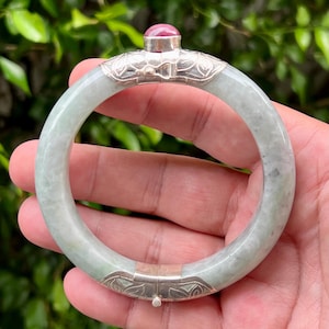 May include: A light green jade bangle bracelet with silver accents and a small, round, red gemstone. The silver sections feature decorative floral patterns. The bracelet is held in a hand against a blurred green background.
