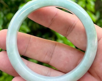 Op de afbeelding: Een gladde, cirkelvormige armband gemaakt van doorschijnend lichtgroen jadeiet. De armband heeft een gepolijst, afgerond oppervlak met subtiele variaties in kleurintensiteit, variërend van zacht muntgroen tot melkachtige wittinten.