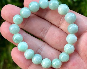 Pode incluir: Uma pulseira de contas verde claro. A pulseira é feita de contas redondas e polidas em diferentes tons de verde claro. As contas são enfiadas para formar um círculo completo. A pulseira é segurada em uma mão.