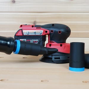 Puede incluir: Una lijadora inalámbrica roja y negra con una manguera negra y dos accesorios negros con detalles azules. La lijadora tiene una batería. El fondo es una superficie de madera marrón claro.