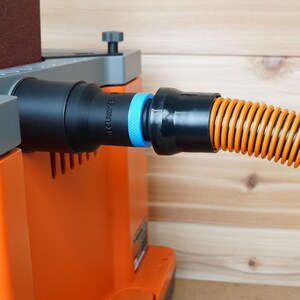 May include: A close-up of a black dust collection hose connected to a sander. The hose is orange and black with a blue connector. The sander is orange and grey with a brown sanding pad.