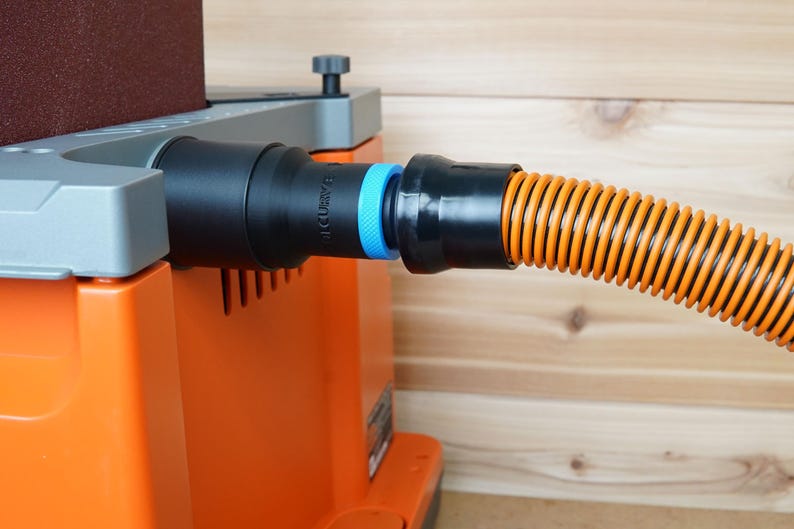 May include: A close-up of a black dust collection hose connected to a sander. The hose is orange and black with a blue connector. The sander is orange and grey with a brown sanding pad.
