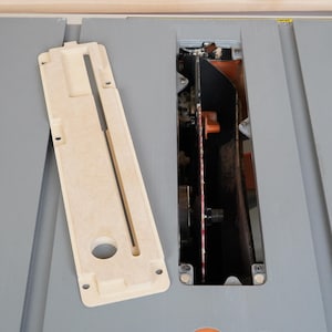 May include: A beige table saw insert and a gray table saw with a blade guard and a blade. The insert is rectangular with a long slot and a circular hole. The saw has a black blade and orange accents.
