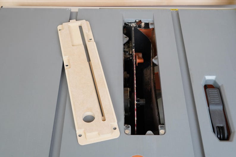 May include: A beige table saw insert and a gray table saw with a blade guard and a blade. The insert is rectangular with a long slot and a circular hole. The saw has a black blade and orange accents.