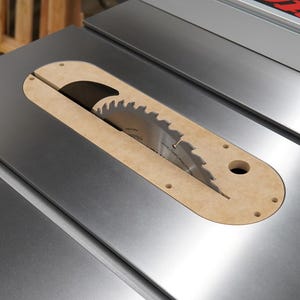May include: Close-up of a table saw with a silver metal surface. A circular saw blade is visible, surrounded by a tan-colored insert. The saw blade has sharp teeth. The saw is designed for woodworking and cutting materials.