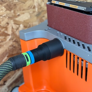 May include: Close-up of a black dust collection port on an orange and gray belt sander. A black hose with green and black braided covering is connected to the port. The hose has a blue and green ring around it. The text "QUICK-RELEASE" is visible on the hose.