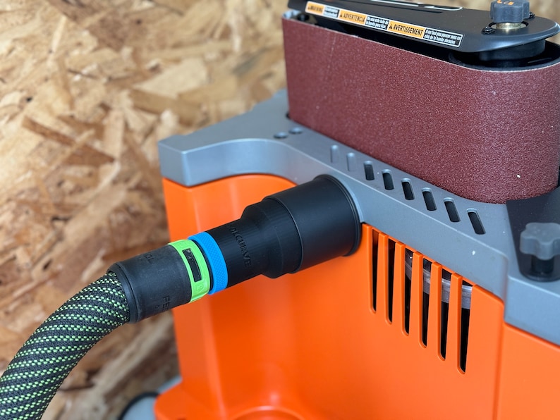 May include: Close-up of a black dust collection port on an orange and gray belt sander. A black hose with green and black braided covering is connected to the port. The hose has a blue and green ring around it. The text "QUICK-RELEASE" is visible on the hose.