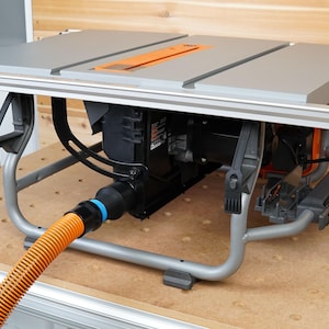 Op de afbeelding: Een grijze tafelzaag met een oranje zaagblad en een zwarte motor. De zaag staat op een metalen onderstel met een stofzuiger aansluiting en een slang die eraan vastzit. De zaag staat op een houten oppervlak.