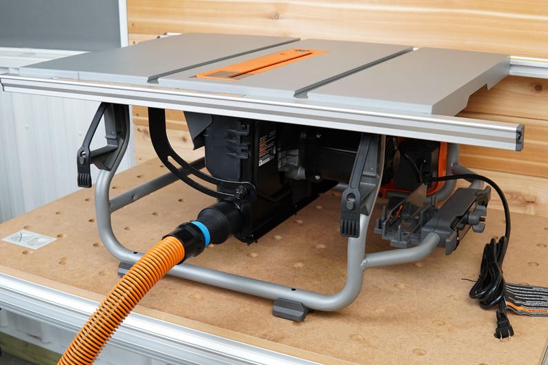 Op de afbeelding: Een grijze tafelzaag met een oranje zaagblad en een zwarte motor. De zaag staat op een metalen onderstel met een stofzuiger aansluiting en een slang die eraan vastzit. De zaag staat op een houten oppervlak.