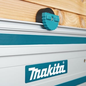 Op de afbeelding: Een close-up van een zilveren en teal Makita railsäge met een zwarte en teal instelknop. De railsäge is gemonteerd op een houten oppervlak.