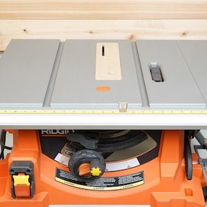 May include: An orange and gray table saw with a silver table top and a yellow measuring tape. The saw has a black control knob and the brand name "RIDGID" is visible on the front.