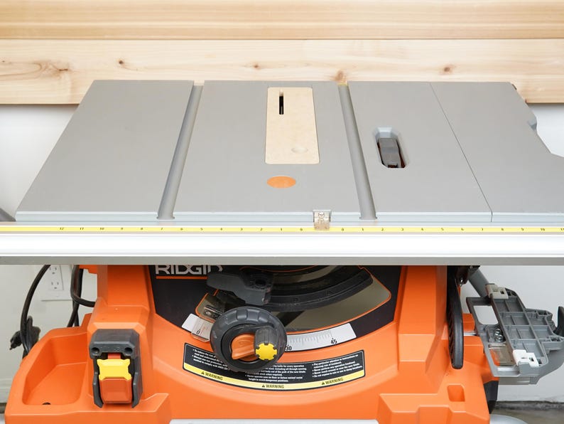 May include: An orange and gray table saw with a silver table top and a yellow measuring tape. The saw has a black control knob and the brand name "RIDGID" is visible on the front.
