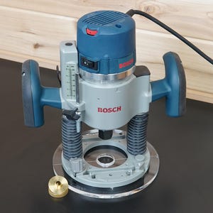 May include: A blue and gray Bosch plunge router featuring two ergonomic handles, a depth adjustment scale, and a black power cord. A small, circular brass guide bushing is positioned on the base plate next to the tool's clear circular sub-base.