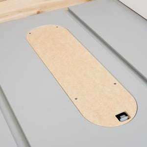 May include: A light gray table saw with a natural wood-toned insert. The insert is an elongated oval shape and is secured with small screws. The saw has a smooth, clean surface.