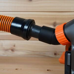 May include: Close-up of a black and orange power tool with a dust collection hose attached. The tool has a black body with an orange dust port and a black hose with an orange corrugated section. The tool has the word "BRUSHLESS" printed on it.