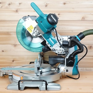 May include: A teal and silver miter saw with a clear blade guard and a black motor housing. The saw has a black handle and a green and black hose attached. The brand name "Makita" is visible on the saw.