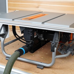 Puede incluir: Una sierra de mesa gris con un motor negro y una mesa de trabajo gris. La sierra est&aacute; sobre un soporte met&aacute;lico con un cable de alimentaci&oacute;n negro y una manguera de recolecci&oacute;n de polvo verde y negra.