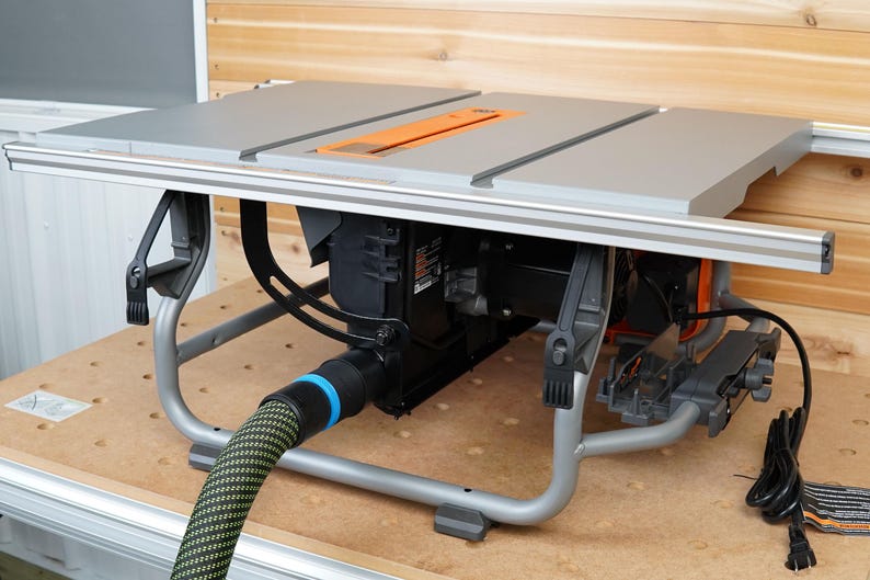 Puede incluir: Una sierra de mesa gris con un motor negro y una mesa de trabajo gris. La sierra est&aacute; sobre un soporte met&aacute;lico con un cable de alimentaci&oacute;n negro y una manguera de recolecci&oacute;n de polvo verde y negra.