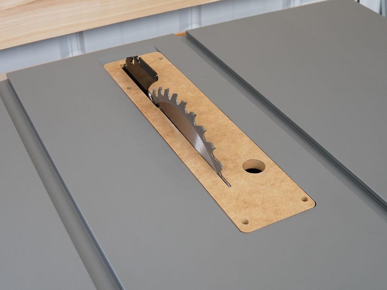 Puede incluir: Primer plano de una hoja de sierra de mesa con una superficie de mesa gris. La hoja es parcialmente visible a trav&eacute;s de una abertura rectangular en la mesa.