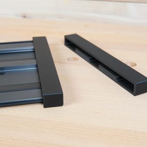 May include: Black metal components, possibly for a tool or fixture, are displayed on a light wood surface. One piece is rectangular with the brand name "RIDGID" visible. The other is a separate, rectangular piece.