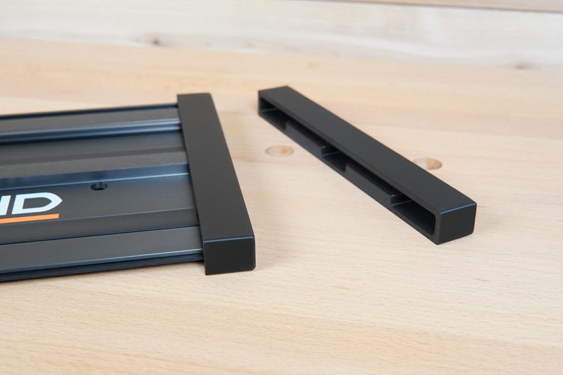 May include: Black metal components, possibly for a tool or fixture, are displayed on a light wood surface. One piece is rectangular with the brand name "RIDGID" visible. The other is a separate, rectangular piece.