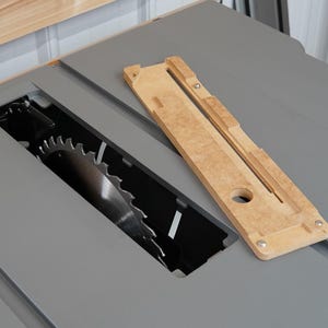 Puede incluir: Primer plano de una sierra de mesa con un inserto de madera para una hoja de dado. El inserto est&aacute; hecho de madera clara y tiene una ranura para que pase la hoja. La sierra de mesa es gris y tiene una hoja negra.