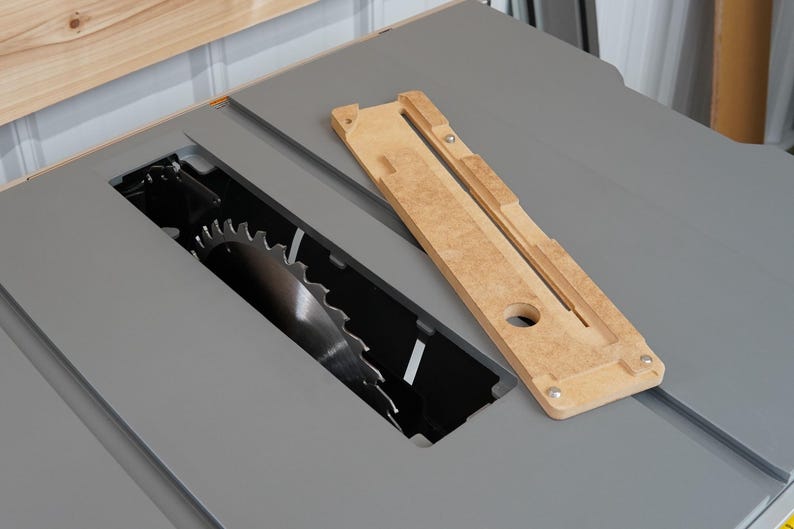 Puede incluir: Primer plano de una sierra de mesa con un inserto de madera para una hoja de dado. El inserto est&aacute; hecho de madera clara y tiene una ranura para que pase la hoja. La sierra de mesa es gris y tiene una hoja negra.