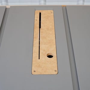 Puede incluir: Primer plano de una mesa de color gris con un recorte rectangular para un elevador de fresadora. El recorte est&aacute; hecho de madera marr&oacute;n claro y tiene un peque&ntilde;o agujero en el centro.